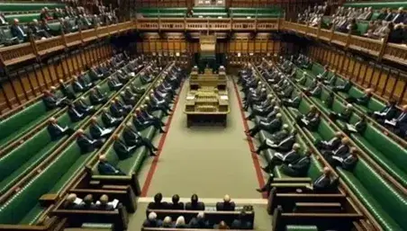 Interior view of the UK House of Commons with green benches, sparsely seated individuals in business attire, and ornate golden ceiling.