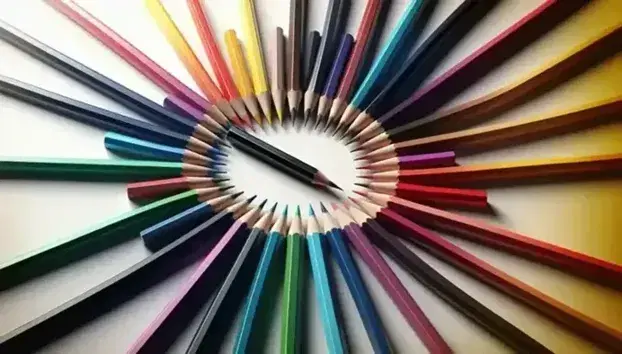 Matite colorate disposte a cerchio con punte convergenti al centro e matita nera più lunga al centro che punta verso l'esterno, su sfondo bianco.
